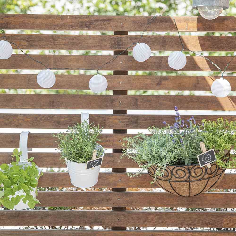 Drewniany płot z zwisającymi roślinami i białymi lampkami. Różne zioła w doniczkach i koszykach tworzą letnią atmosferę.