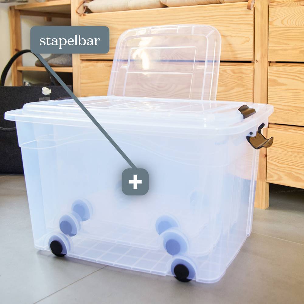 Przezroczysta plastikowa skrzynka na kółkach przed drewnianą komodą. Etykieta wskazuje "możliwość układania w stos", podkreślając jej funkcję efektywnego przechowywania.