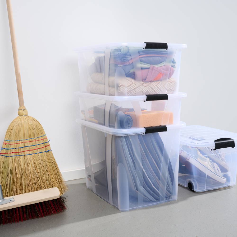 Na podłodze stoją trzy przezroczyste pudła magazynowe ułożone jedno na drugim, wypełnione przedmiotami gospodarstwa domowego. Obok nich, przy ścianie, oparta jest miotła.