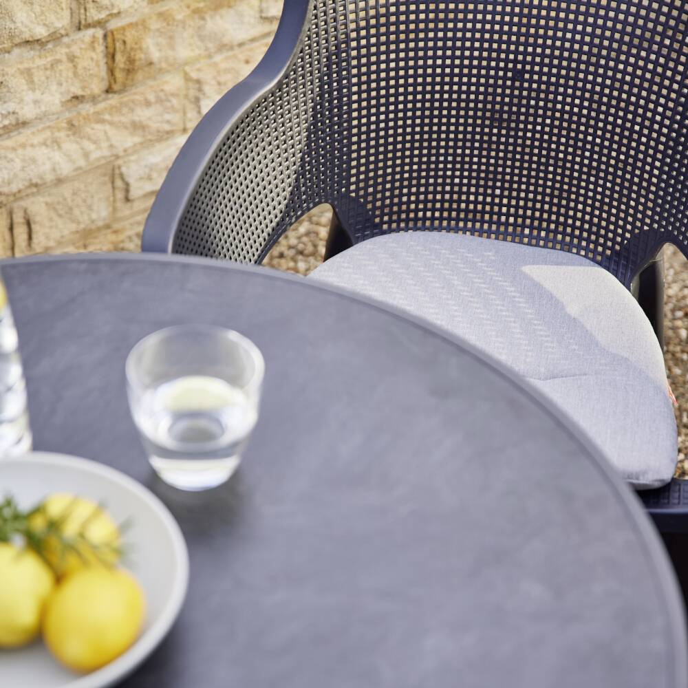 Szklanka wody stoi obok talerza z cytrynami na okrągłym stole, przed plecionym krzesłem na zewnątrz, przy kamiennym murze.