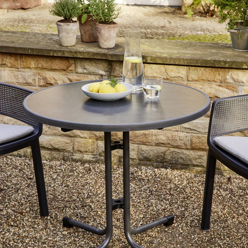 Okrągły stół na dworze z dwoma krzesłami. Na stole stoją dzbanek wody, szklanka i miska z cytrynami. W tle rośliny.