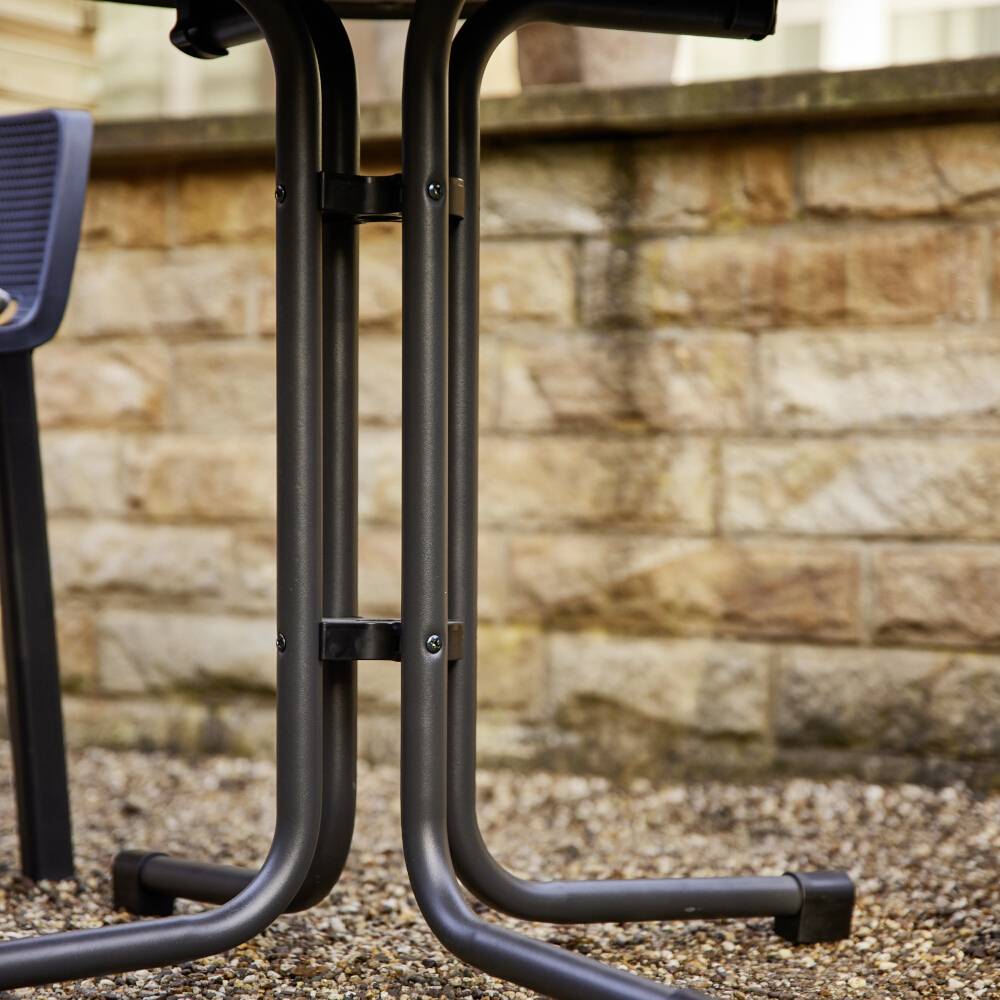 Metalowy stół stoi na żwirowej nawierzchni przed kamiennym murem. Po lewej stronie obrazu znajduje się niebieski fotel.