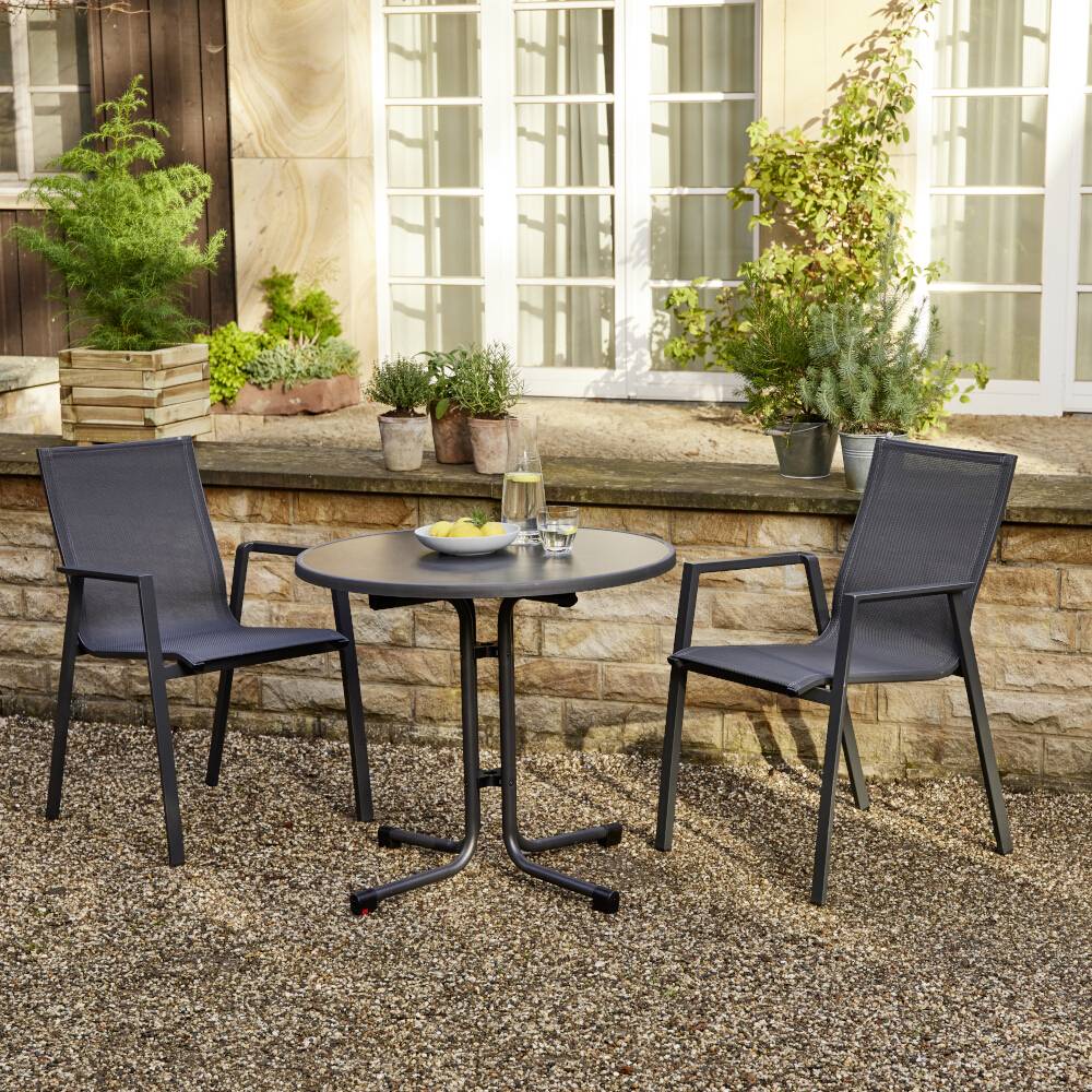 Dwa krzesła i okrągły stół na żwirowej nawierzchni przed niskim murem kamiennym porośniętym roślinami. Na stole stoją szklanki i woda.