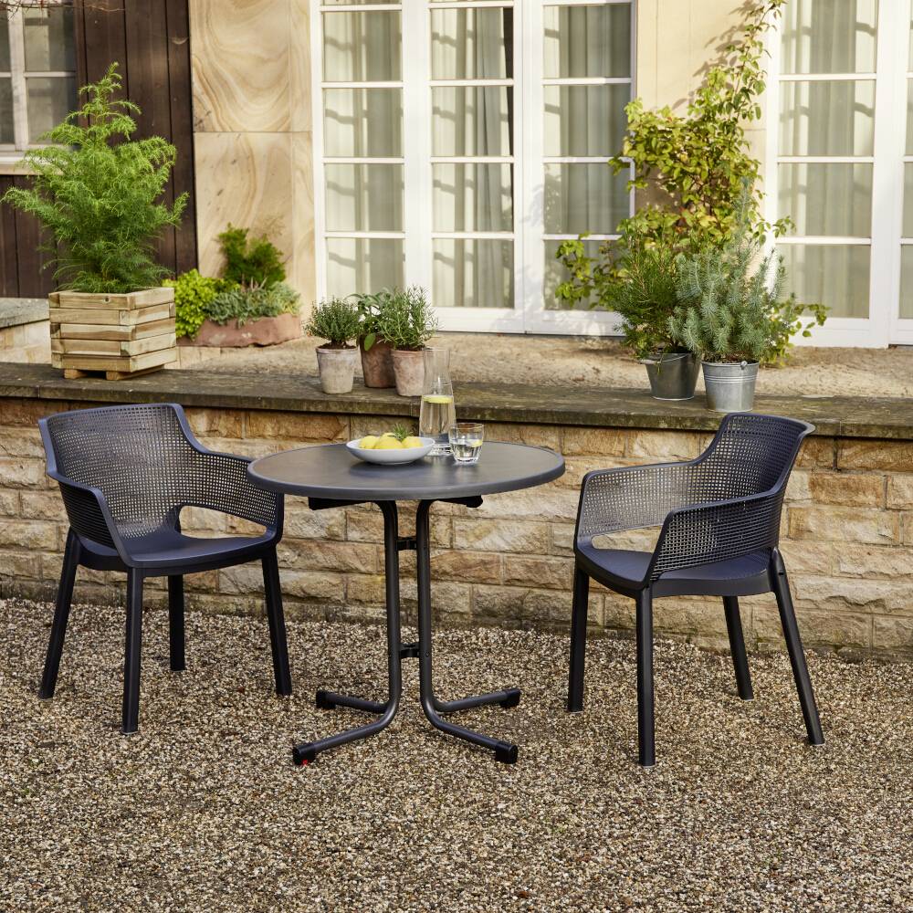 Okrągły stół z dwoma krzesłami stoi na powierzchni żwiru. Na stole znajduje się misa z owocami. W tle: kamienny mur porośnięty roślinami.