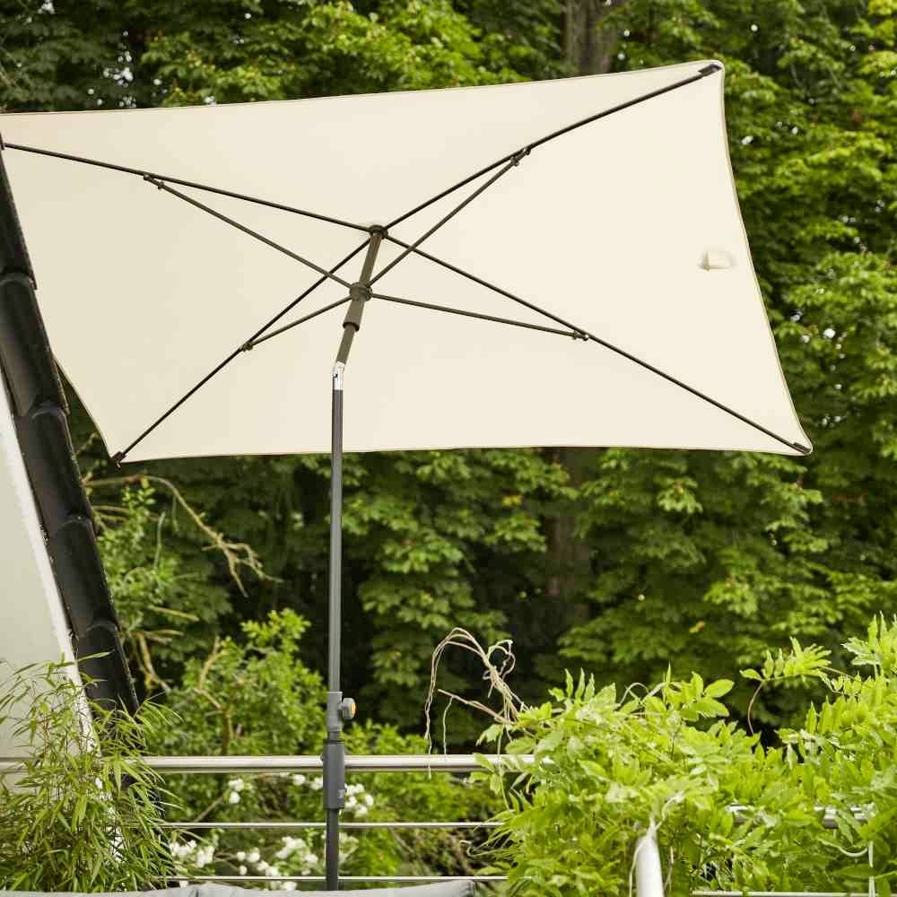 Kremowy parasol na tarasie, na tle zieleni drzew i roślin. Parasol rzuca cień.