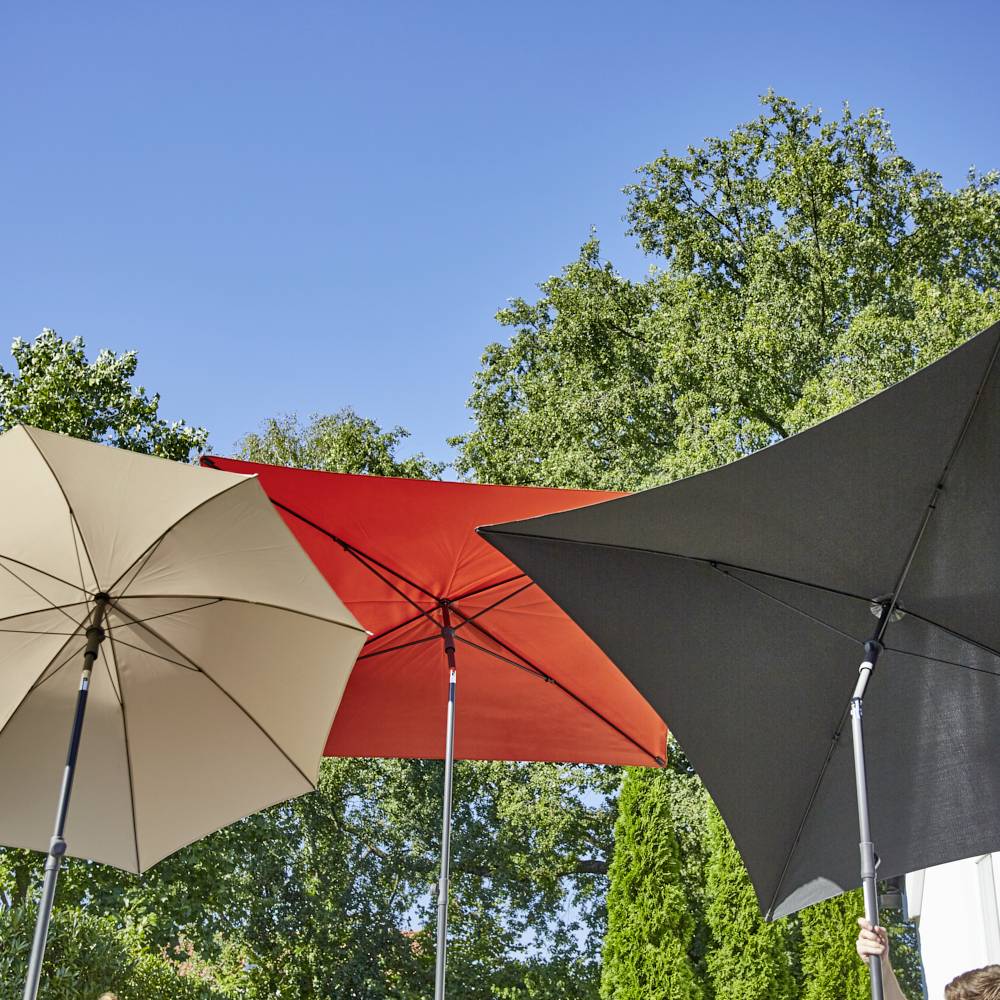 Trzy otwarte parasole przeciwsłoneczne w kolorach beżowym, czerwonym i czarnym stoją przed zielonym tłem z drzewami pod czystym niebem.