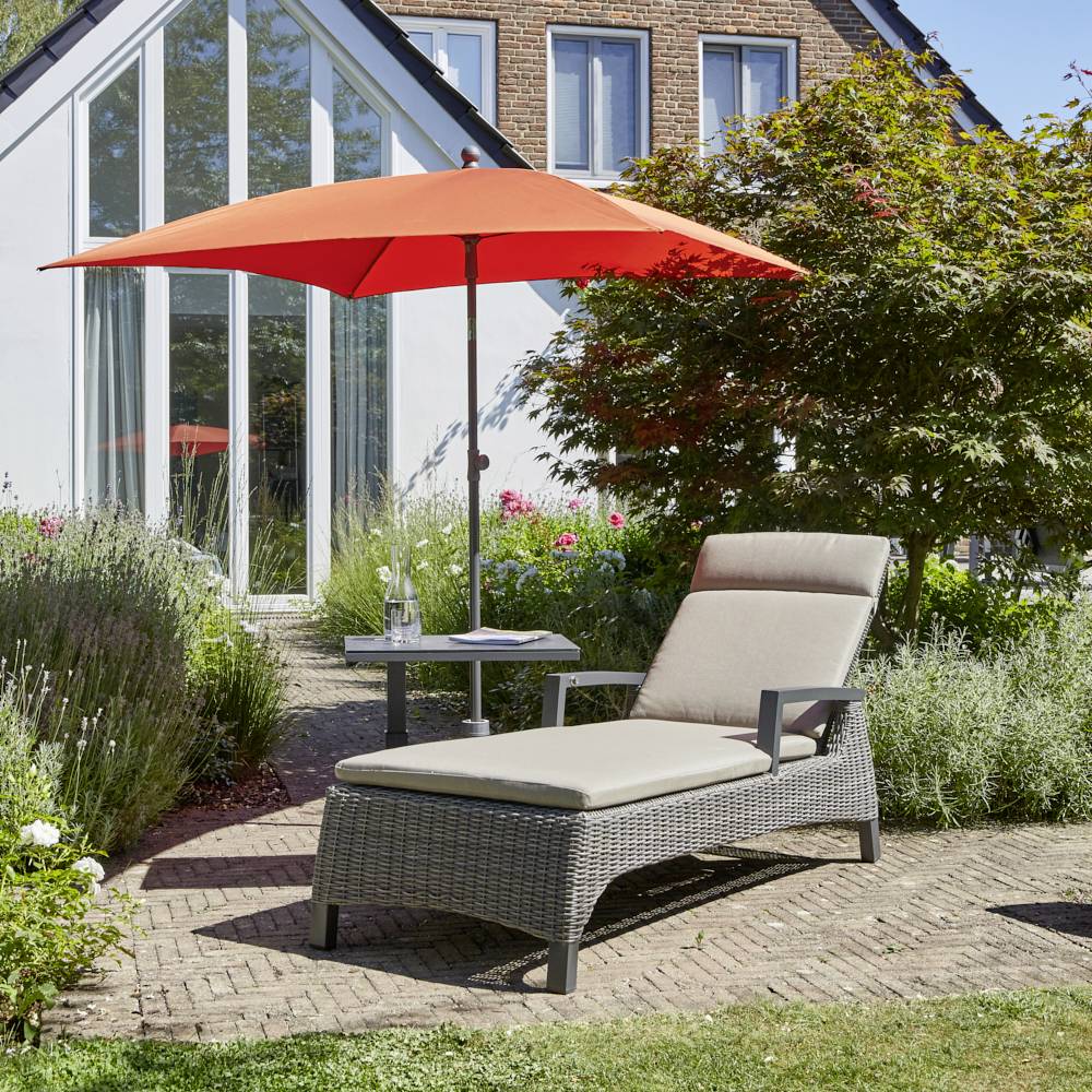 Leżak i parasol przeciwsłoneczny w ogrodzie przed nowoczesnym domem, otoczony roślinami i kwiatami, słoneczny dzień.