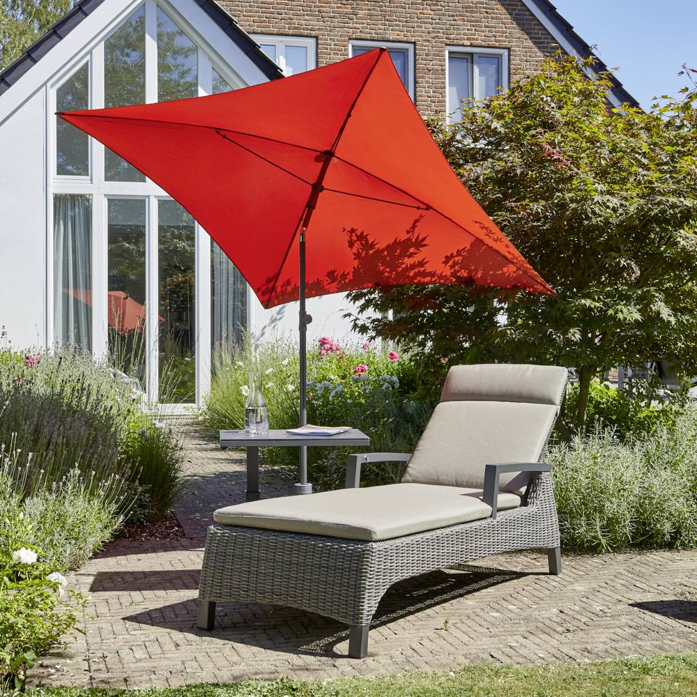 Leżak z poduszką i czerwonym parasolem przeciwsłonecznym w ogrodzie przed domem. Cienie i kwitnące rośliny w tle.