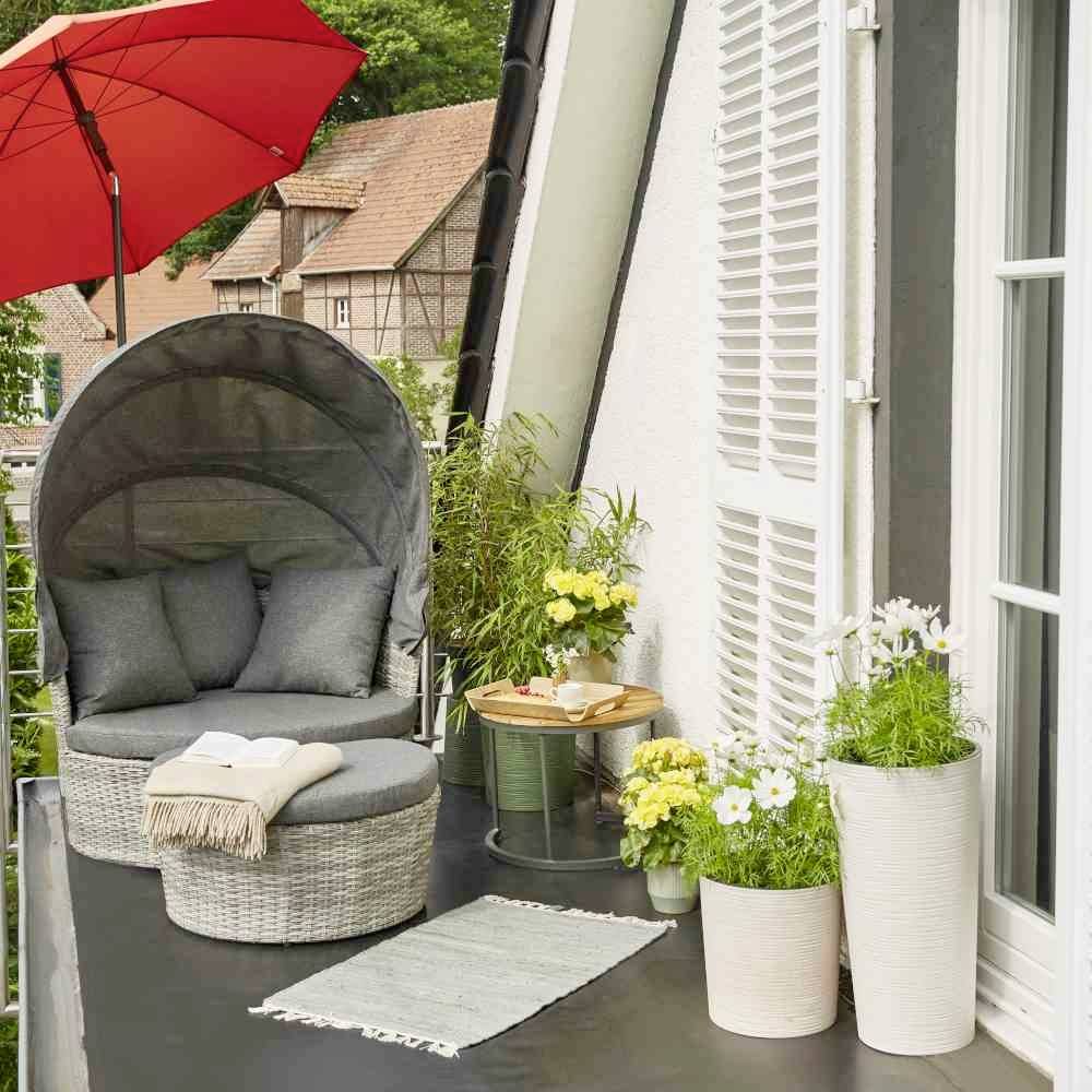 Przytulny balkon z okrągłą ławką, czerwonym parasolem, roślinami w białych doniczkach oraz stolikiem z książką; wiejskie otoczenie.