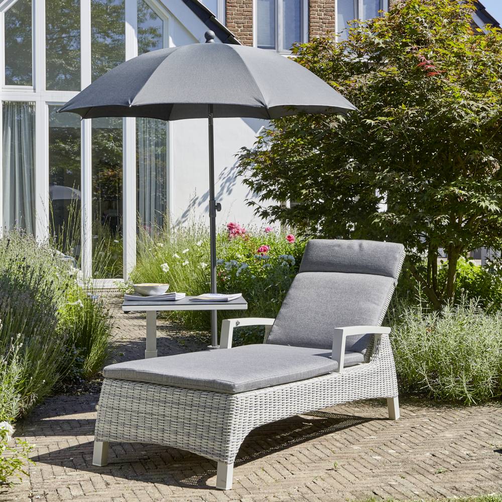 Przytulny obszar ogrodowy z rattanowym leżakiem, szarym parasolem i małym stolikiem na brukowanym tarasie przed nowoczesnym domem ze szkła.
