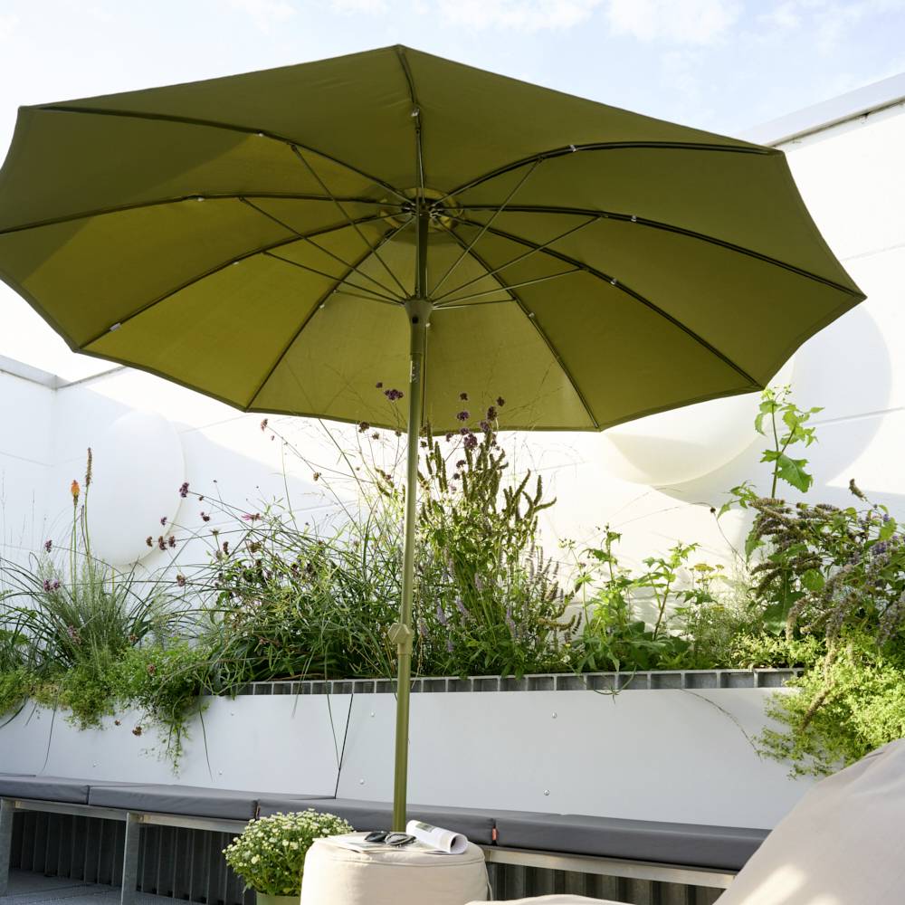 Duży zielony parasol stoi na tarasie, otoczony roślinami. To słoneczny dzień z nielicznymi chmurami na niebie.