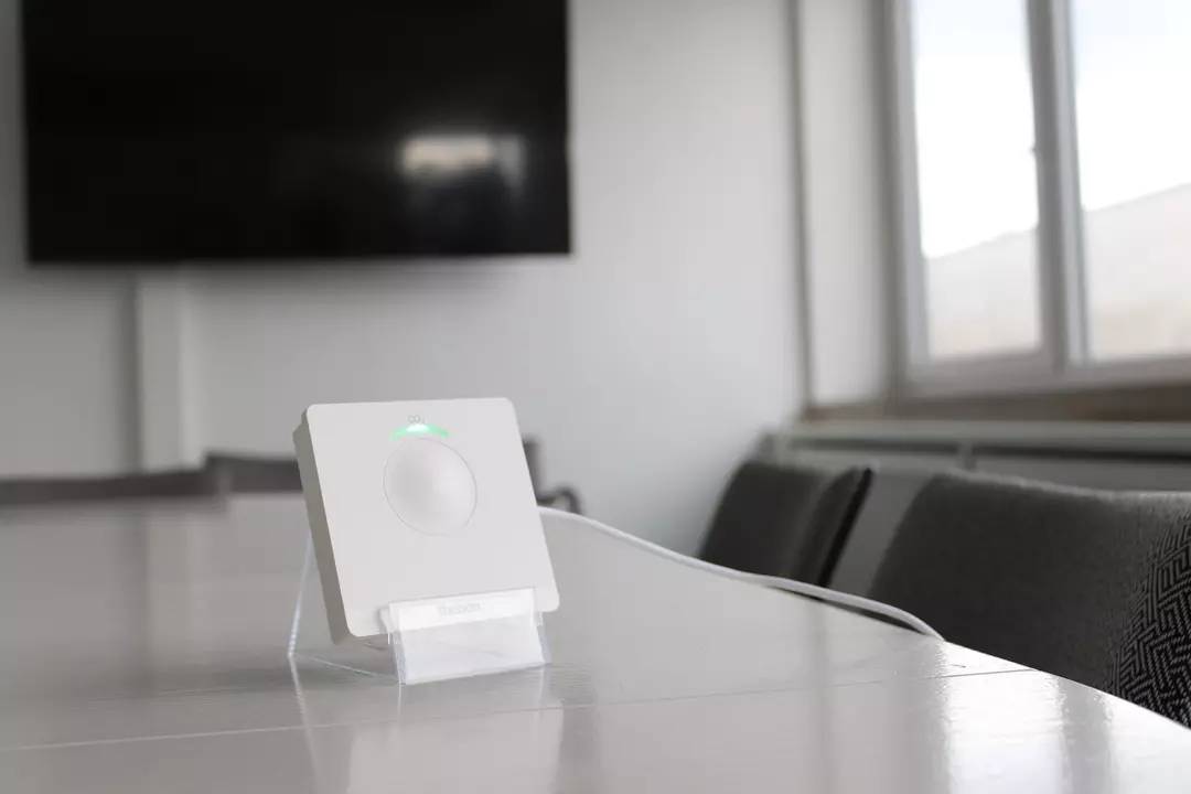 Prostokątne urządzenie z zielonym sygnalizatorem świetlnym stoi na stole w sali konferencyjnej, na tle ekranu.