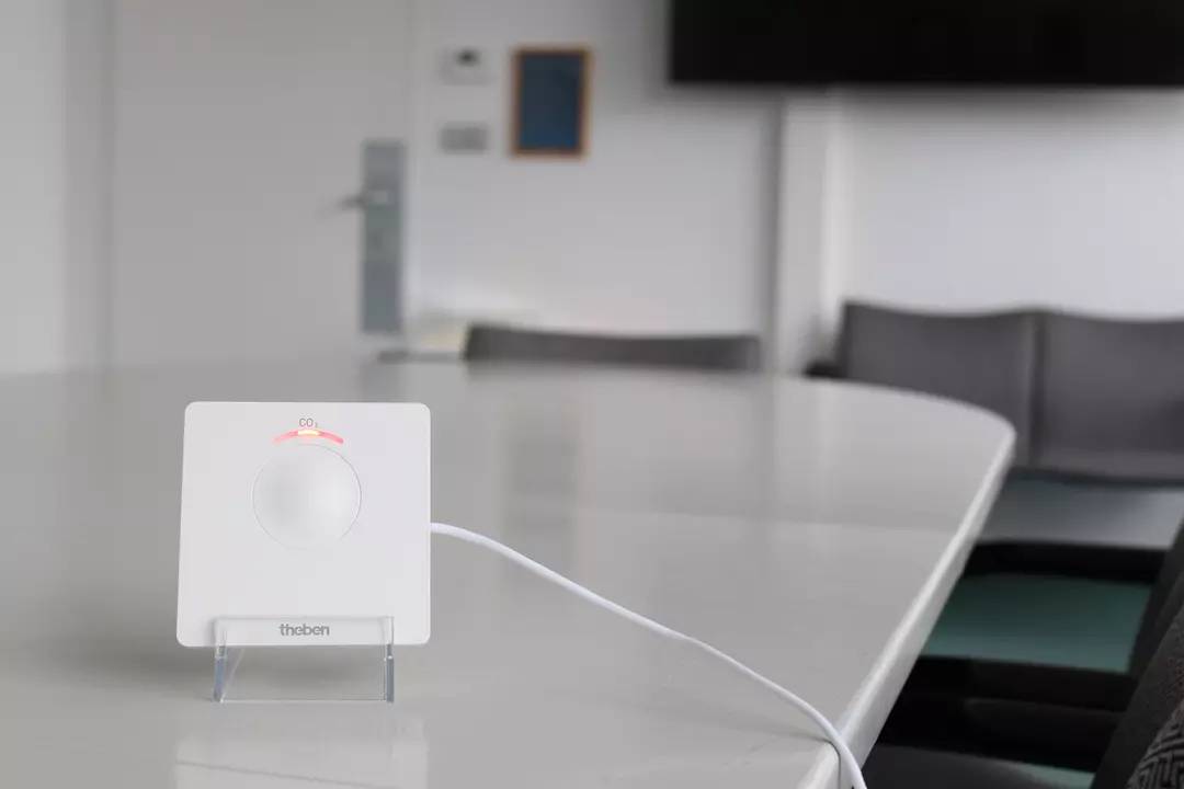 Biały regulator termostatu leżący na stole w biurze. Regulator wskazuje ustawienie 'Komfort'.