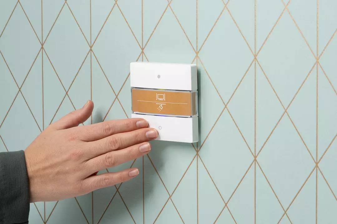 Dłoń dotyka nowoczesnego włącznika światła na jasnoniebieskiej ścianie ze złotymi liniami. Włącznik posiada graficzny wyświetlacz.