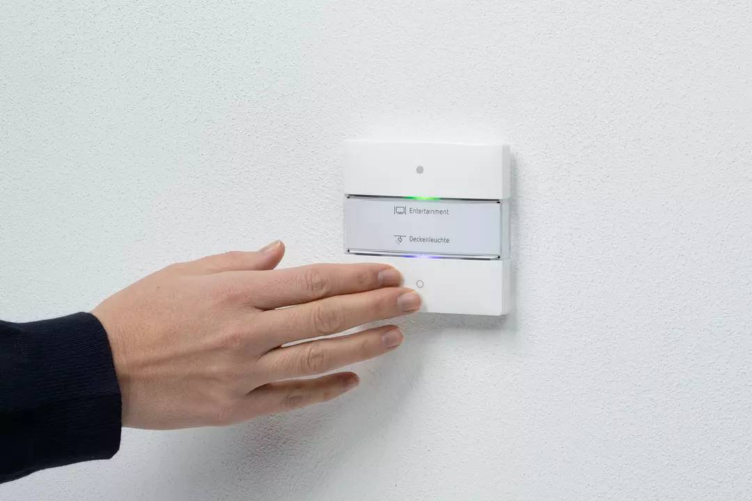 Dłoń dotyka włącznika na ścianie. Przełącznik ma dwa przyciski, oznaczone jako 'Usuń' i 'Lampa sufitowa'.