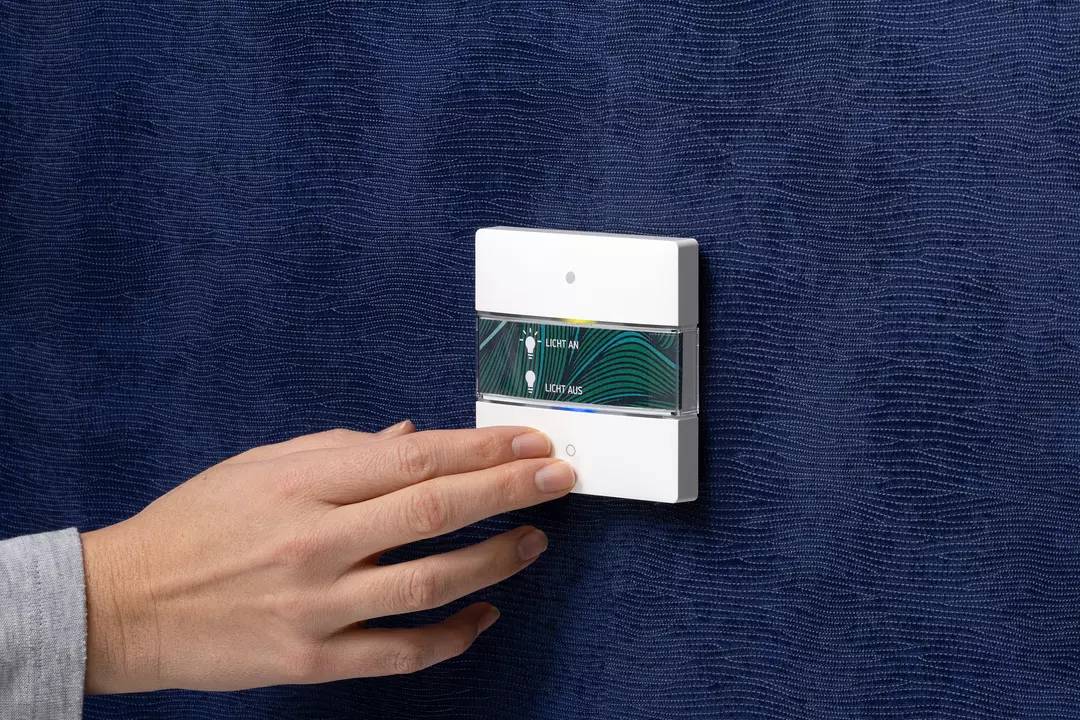 Dłoń dotyka włącznika światła na ścianie. Przełącznik posiada dwa przyciski: 'Włącz światło' i 'Wyłącz światło', w nowoczesnym designie.