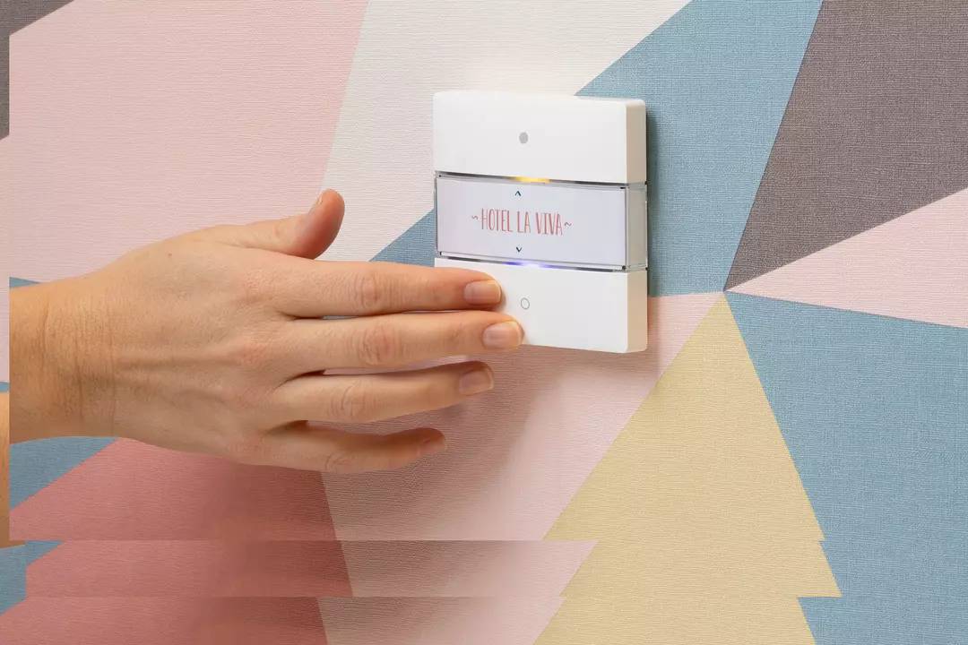 Dłoń zbliża się do nowoczesnego włącznika światła na ścianie o geometrycznym wzorze z napisem 'Hotel La Viva'.