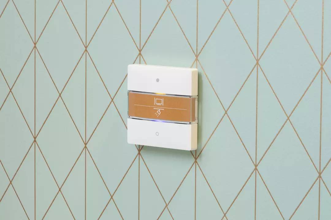 Biały, kwadratowy włącznik światła z brązowym paskiem pośrodku. Przełącznik jest zamontowany na ścianie w kolorze miętowej zieleni.