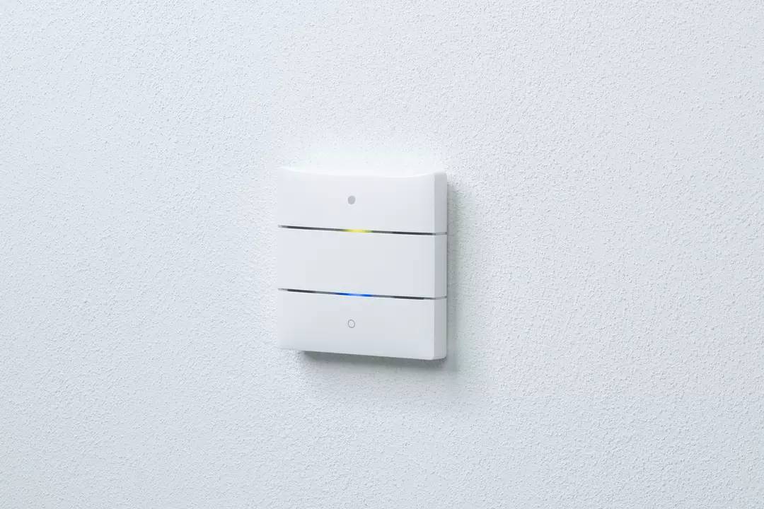 Biały włącznik światła z trzema poziomymi paskami na białej ścianie. Mała żółta i niebieska dioda LED świecą na urządzeniu.