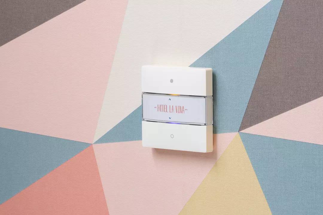 Włącznik światła na ścianie z geometrycznym wzorem. Przełącznik jest oznaczony napisem 'Hotel la Viva'.