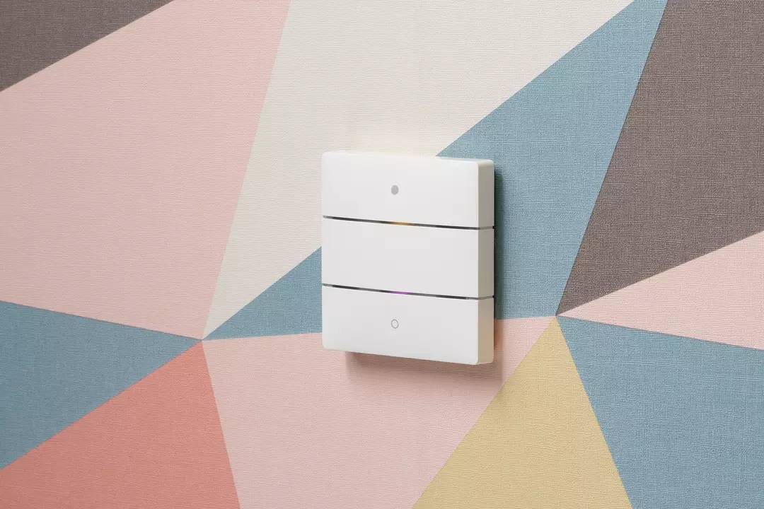 Biały, prostokątny włącznik światła z trzema poziomymi liniami zamontowany na kolorowej, geometrycznie wzorzystej ścianie.