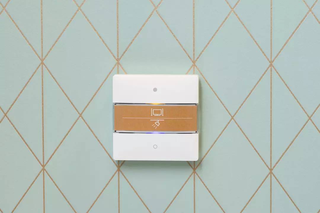 Biały włącznik światła na jasnoniebieskiej ścianie w geometryczny wzór rombu. Przełącznik posiada symbol światła.