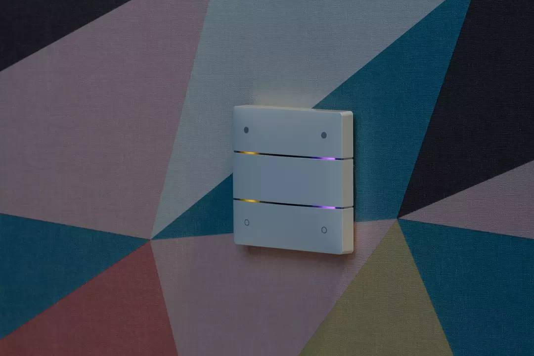 Trójczęściowy włącznik światła na kolorowej, geometrycznej tapecie. Przełącznik posiada świecące kontrolki i czyste linie.