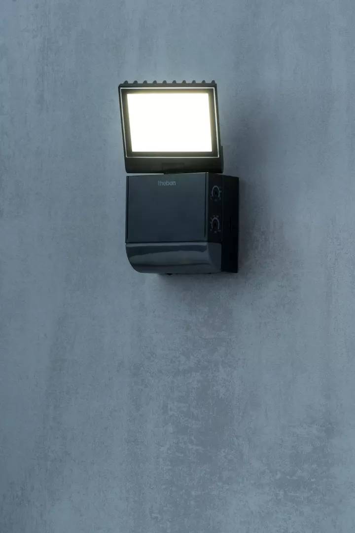 Lampa zewnętrzna na szarej ścianie, włączona, emitująca jasne światło. Odpowiednia do oświetlenia wejść lub ogrodów.