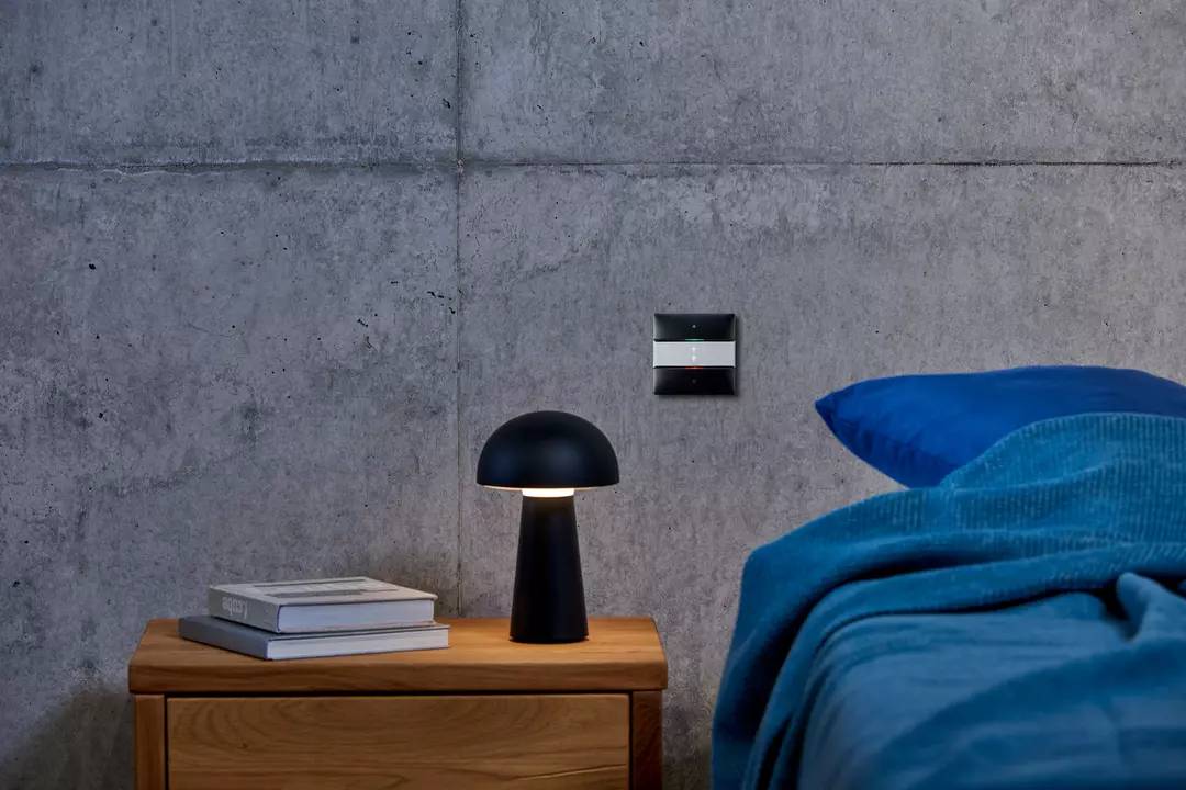 Stolik nocny z lampą i książkami stoi obok łóżka przykrytego niebieską kołdrą, przy szarej ścianie.