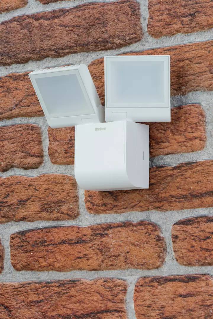 Biały czujnik ruchu jest zamontowany na ścianie z cegły. Detektor posiada dwa prostokątne sensory do wykrywania ruchu.