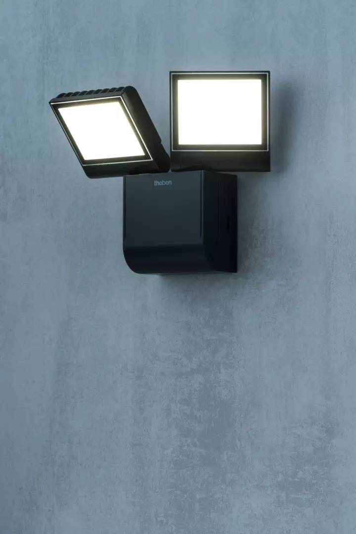 Ciemnoszara lampa zewnętrzna z dwoma kwadratowymi lampami LED, zamontowana na betonowej ścianie, świeci jasno.