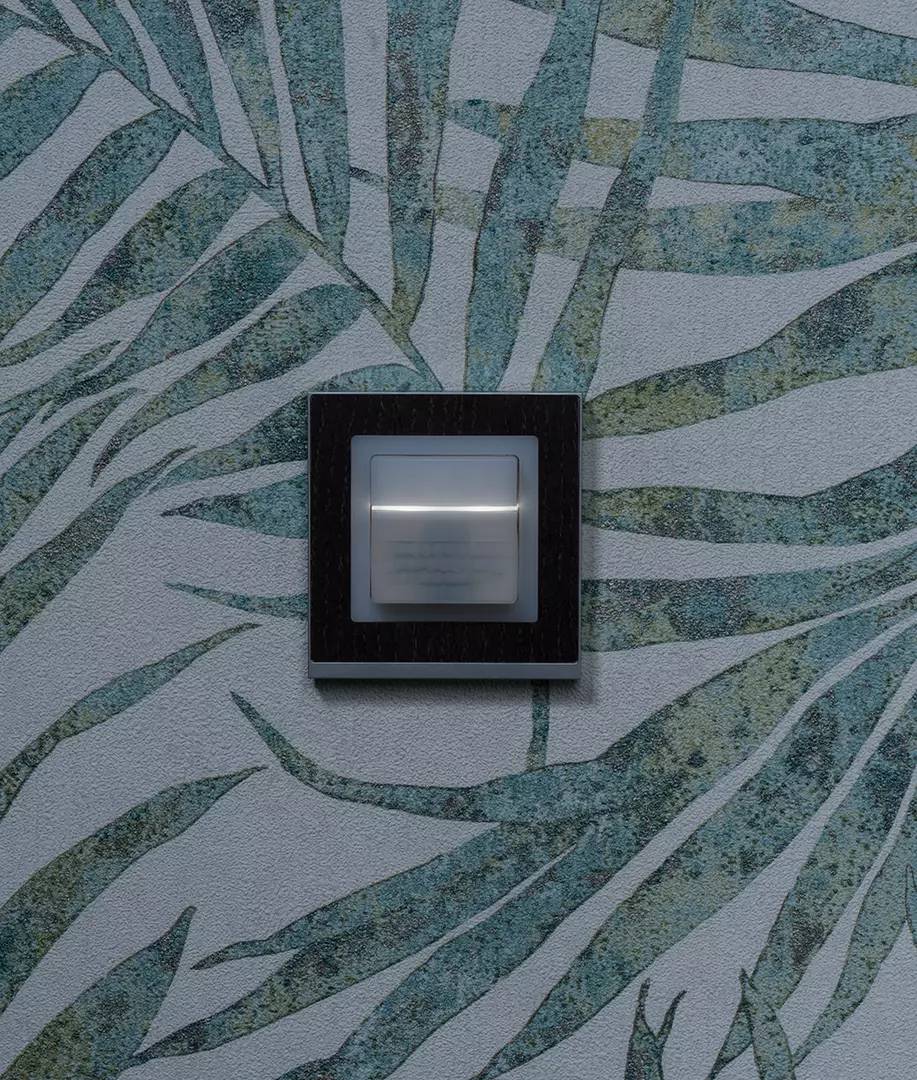 Włącznik światła na ścianie z zielono-szarym wzorem liściastym.