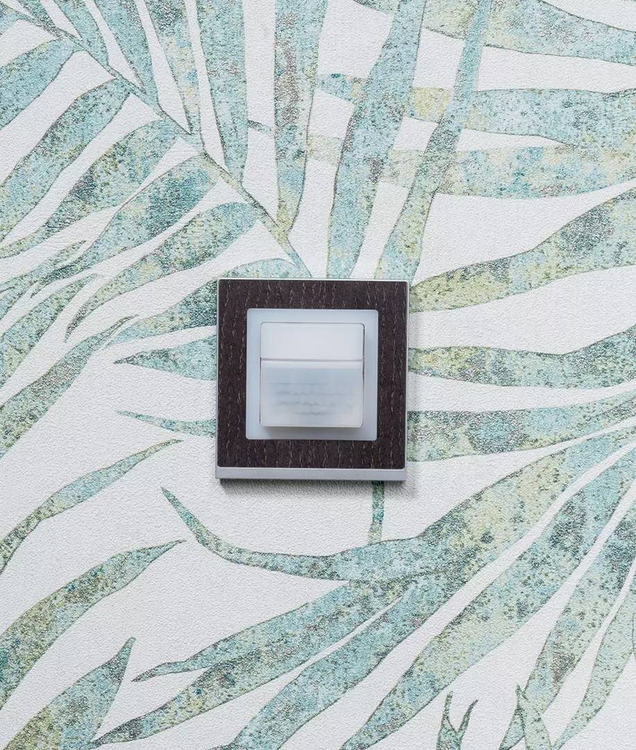 Włącznik światła na tapecie w kwiatowy wzór z liściastymi kształtami w odcieniach zieleni i błękitu na białym tle.