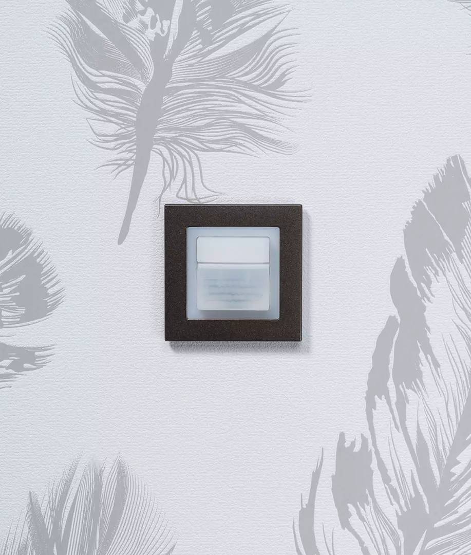 Nowoczesny włącznik światła na ścianie z szarym wzorem piór. Przełącznik posiada czarną ramkę i białą przycisk.