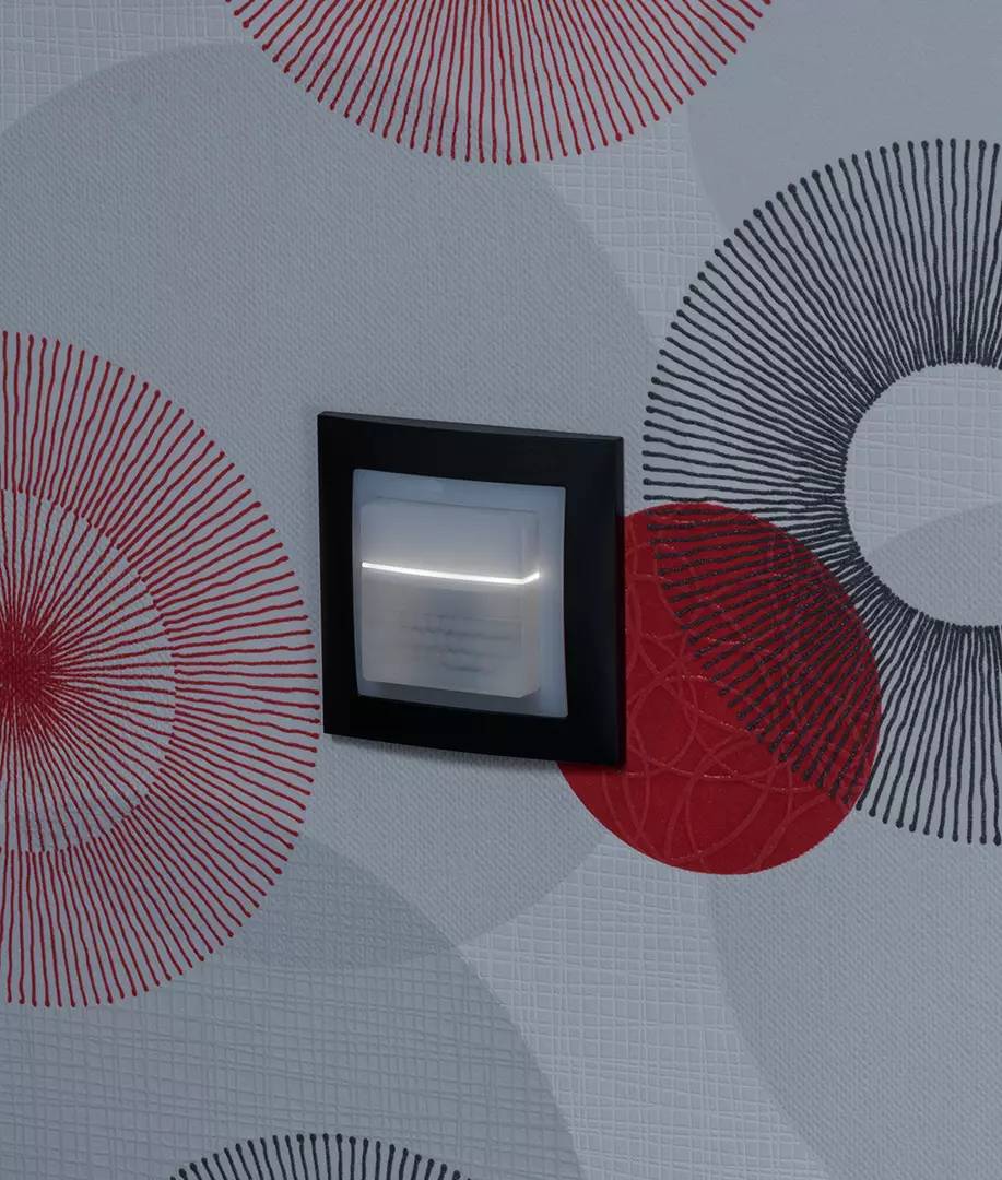 Lampka nocna w czarnej ramce jest zamocowana na ścianie ozdobionej geometrycznym wzorem składającym się z czerwonych i szarych kół.