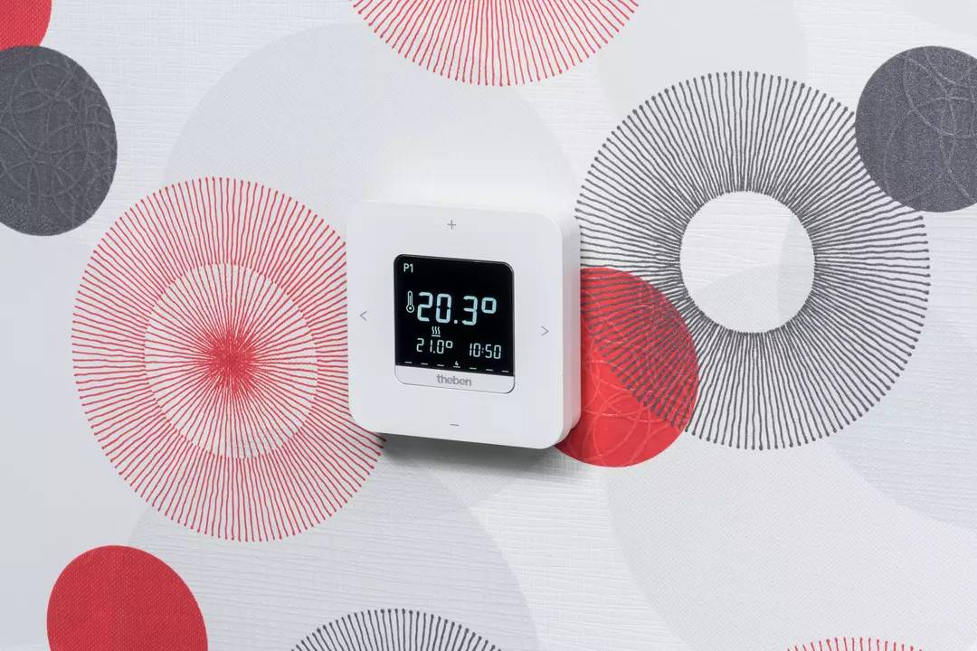 Cyfrowy termostat na ścianie wskazuje aktualną temperaturę 20,3 stopni Celsjusza. Ściana ma wzór w kolorach czerwonym i białym.