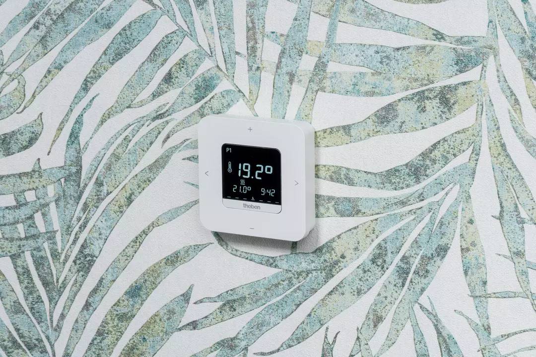 Cyfrowy termostat wyświetla '19,2°C'. Jest zamontowany na wzorzystej ścianie.