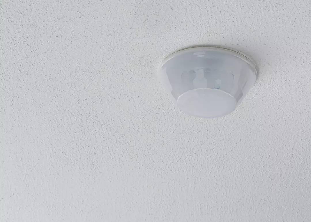 Okrągła, biała lampa sufitowa na białym, strukturalnym suficie. Lampa jest wyłączona i prosta w swoim wzornictwie.