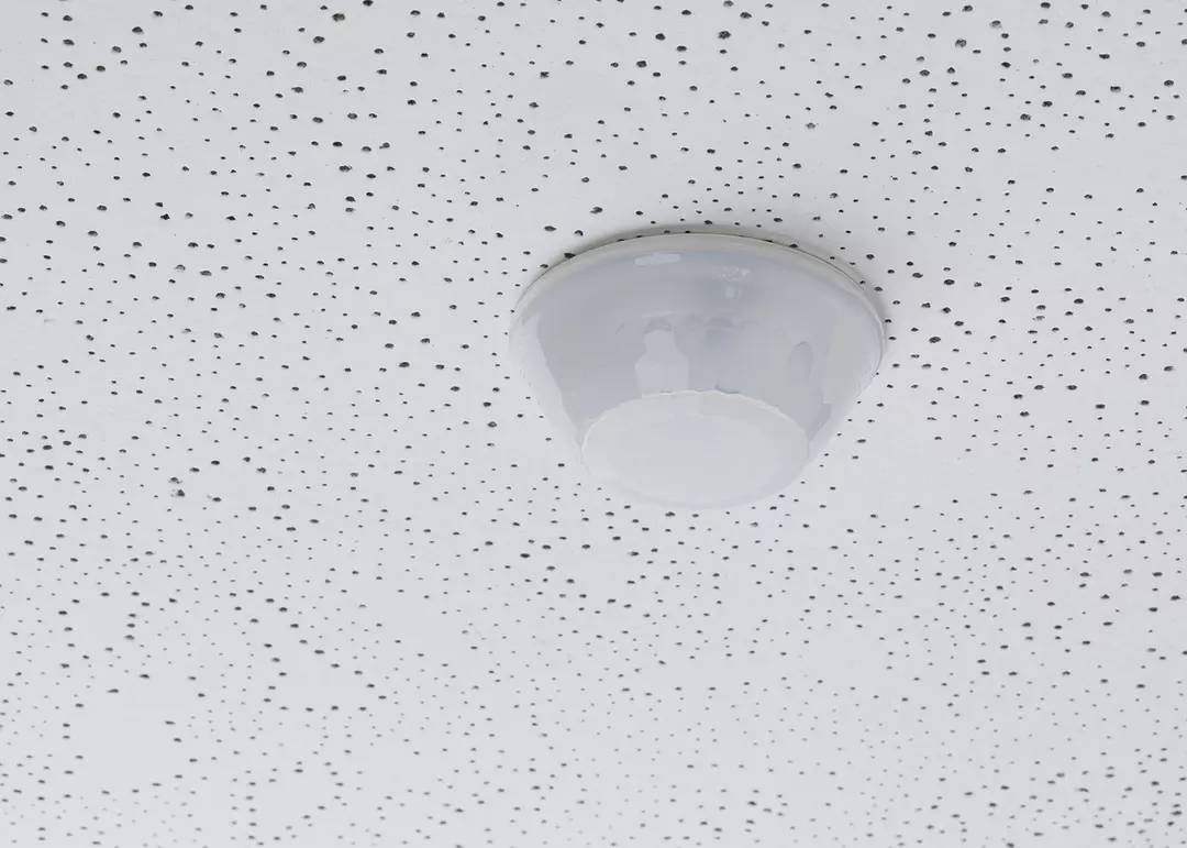Biała lampa sufitowa jest zamontowana na perforowanym suficie i rzuca równomierne światło w pomieszczeniu.