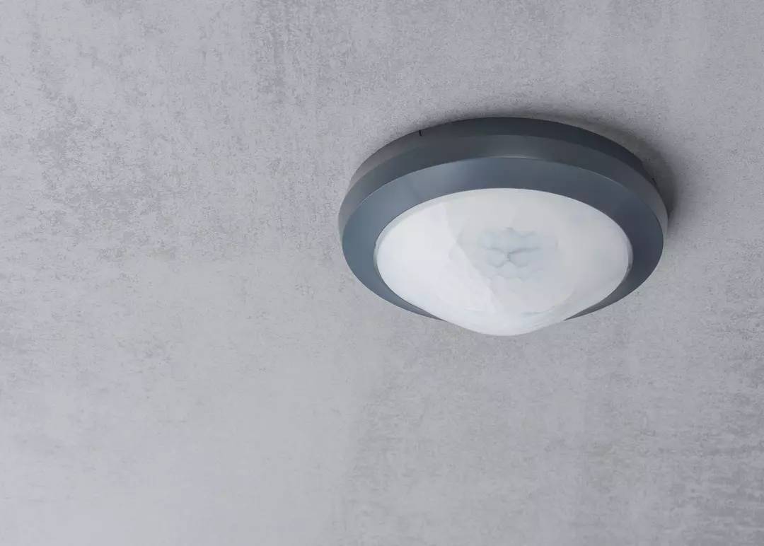 Okrągła lampa zamontowana na suficie, z szarą ramą i białym światłem, na szarym tle.