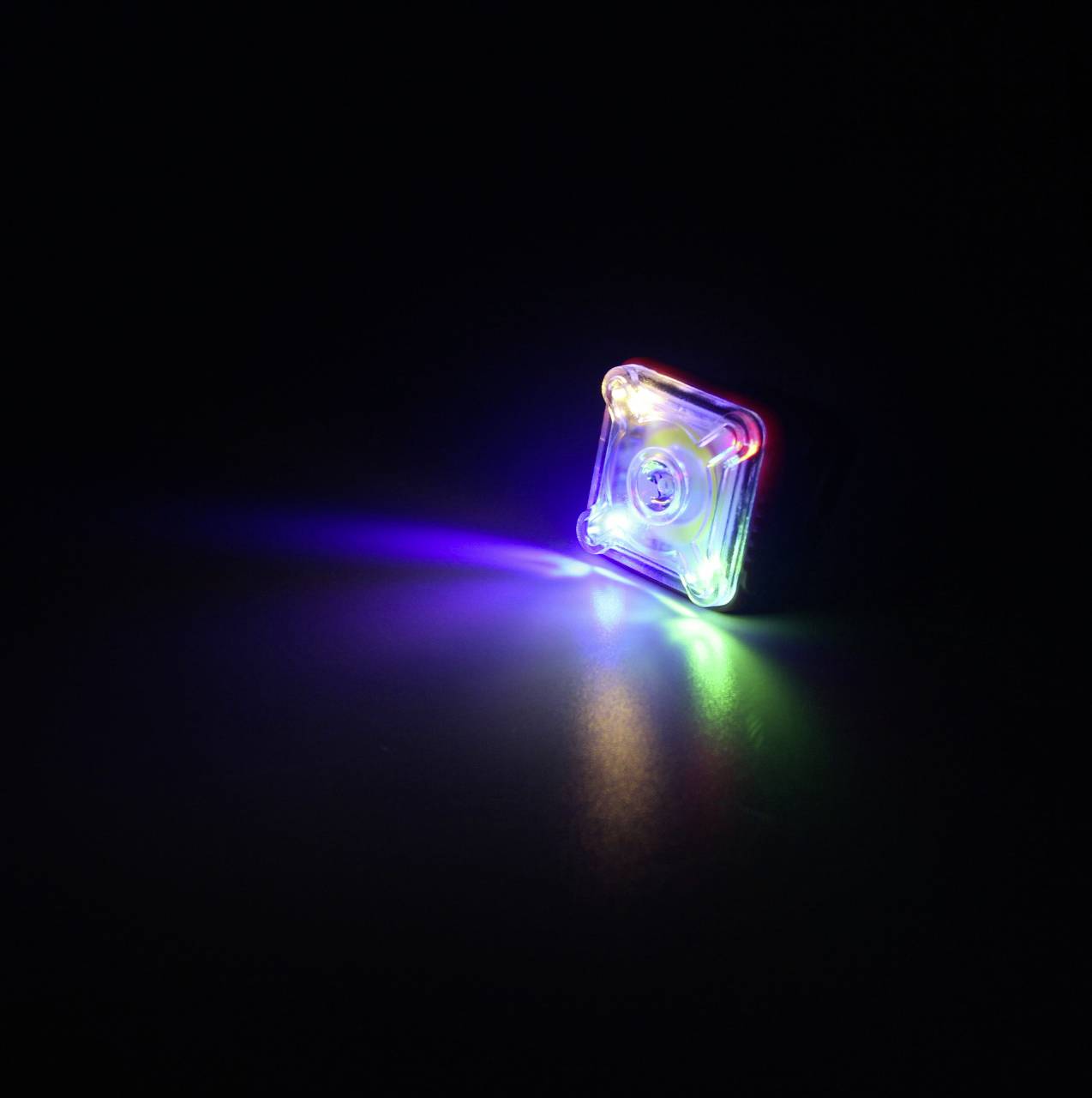 Mały, kwadratowy reflektor LED w różnych kolorach świeci w ciemności. Tworzy on efekt tęczowy na powierzchni.