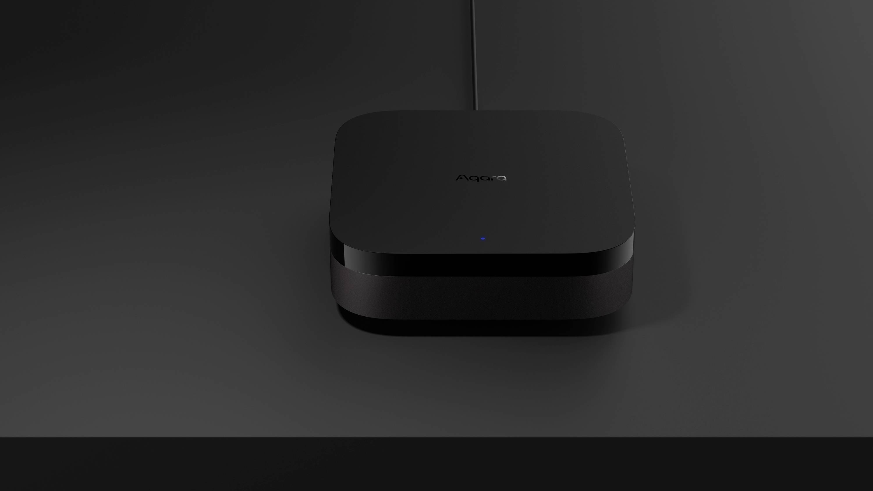 Czarne, kwadratowe urządzenie o zaokrąglonych rogach ze świecącym niebieskim punktem stoi na ciemnym stole.