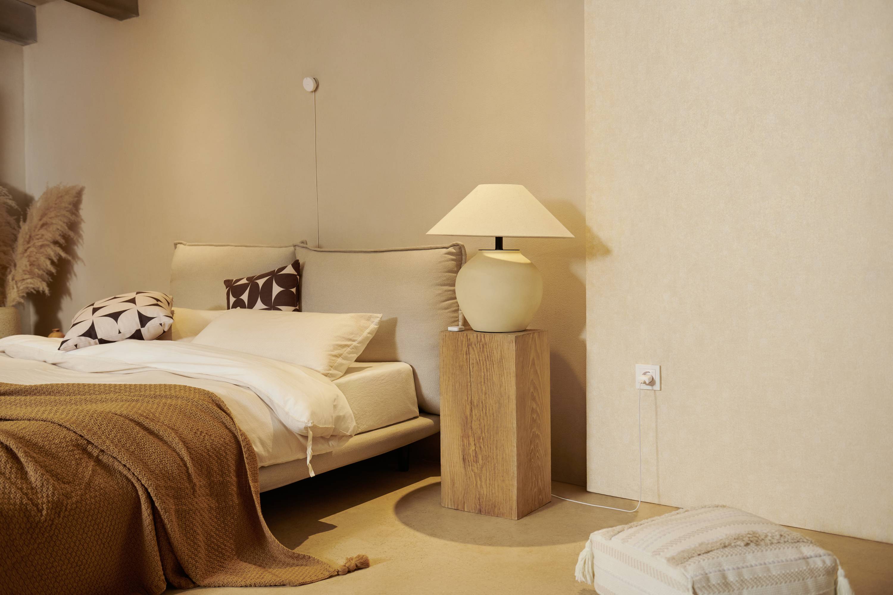 Przytulna sypialnia z podwójnym łóżkiem, beżowymi ścianami, lampką nocną na drewnianym stoliku oraz dekoracyjnymi poduszkami.