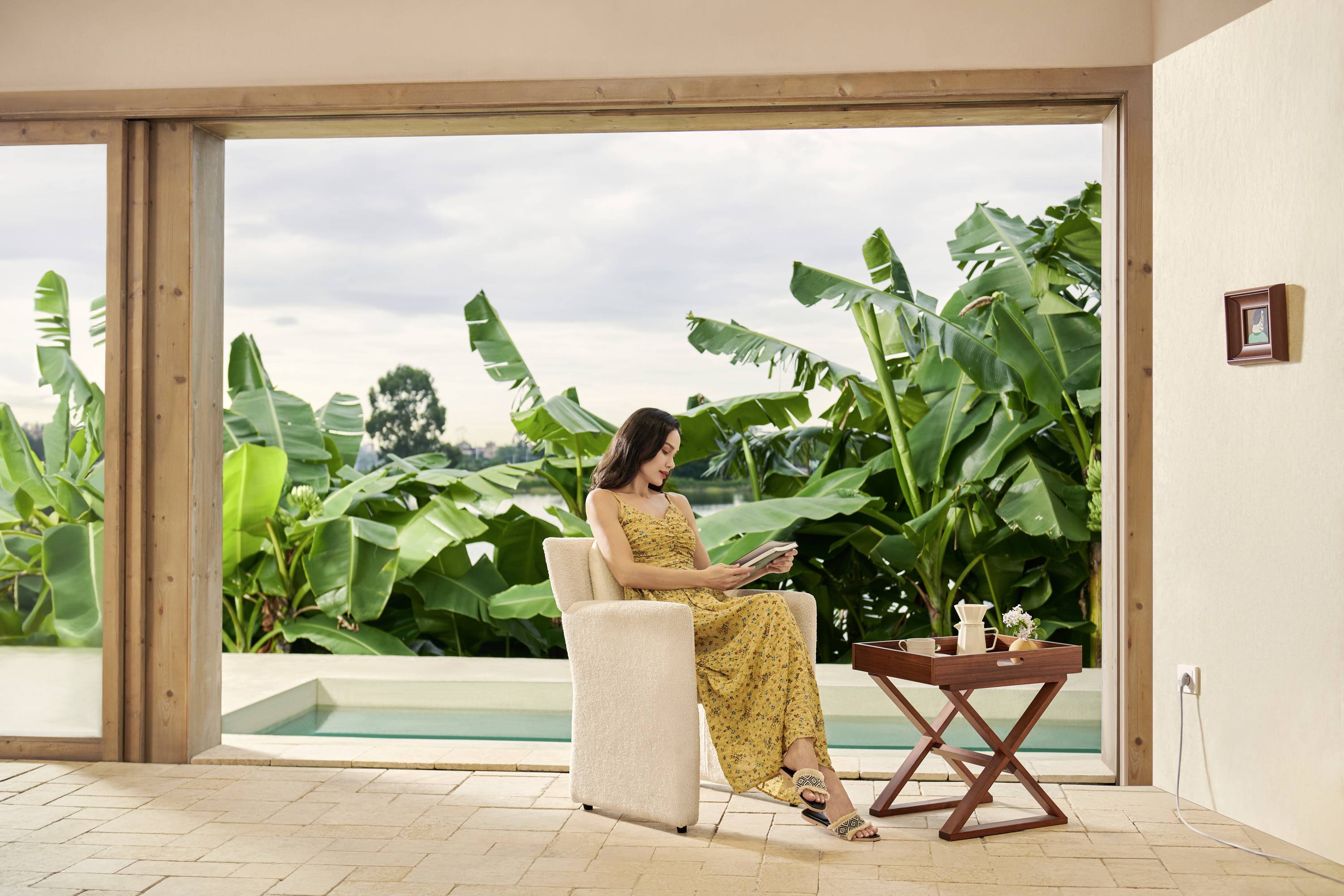 Kobieta w żółtej sukience siedzi w nowoczesnym pomieszczeniu i czyta książkę. W tle widoczne są tropikalne rośliny.