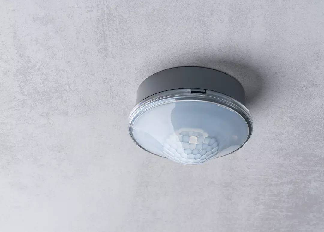 Okrągła lampa sufitowa z nowoczesną, szarą obudową i strukturyzowaną, białą osłoną, zamontowana na szarej ścianie.