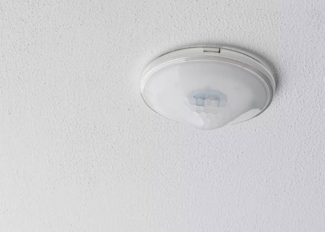 Okrągły, biały czujnik ruchu sufitowy, zamontowany na białym suficie. Główna funkcja: wykrywanie ruchu do sterowania oświetleniem.
