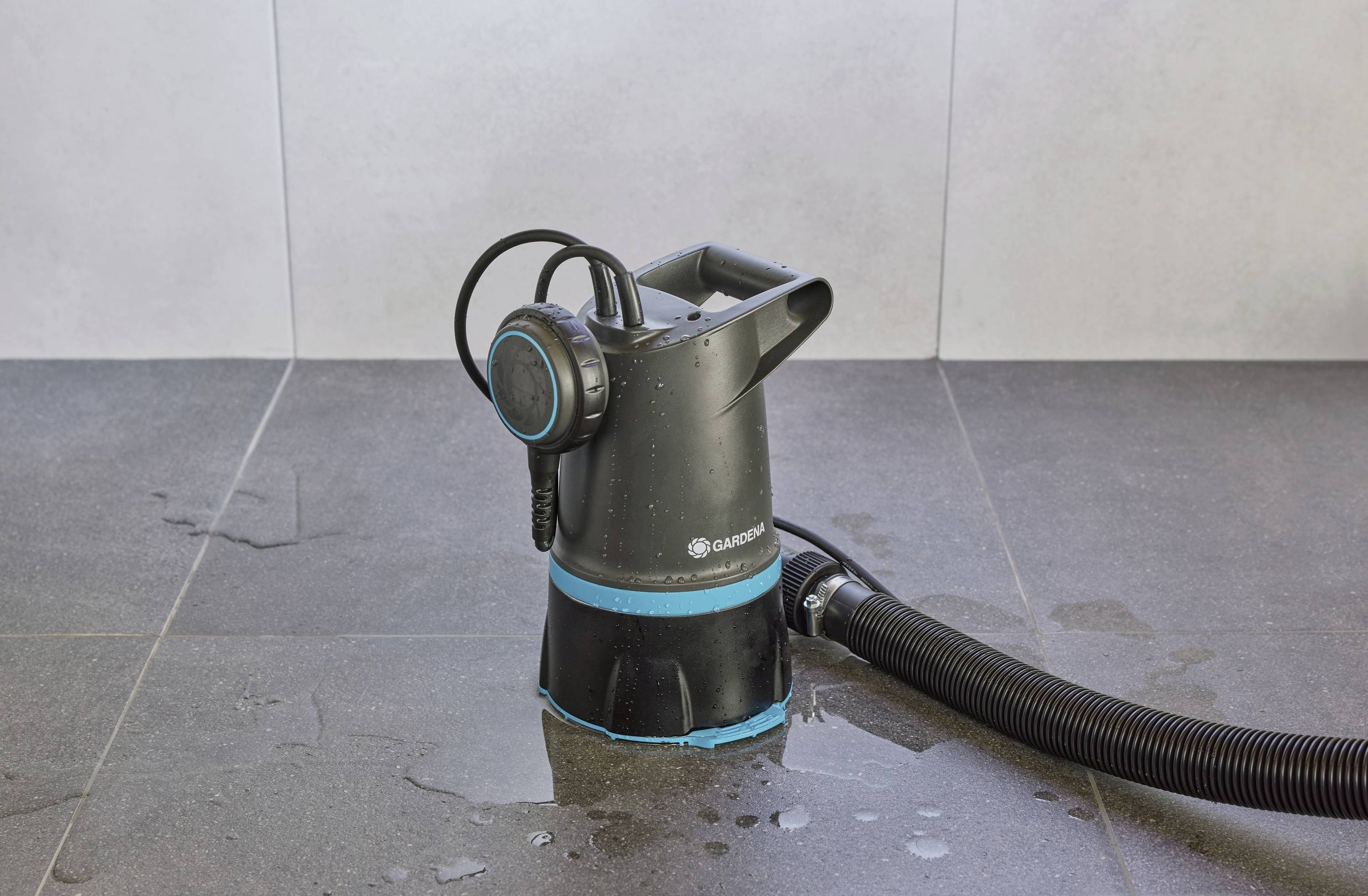 Czarna pompa zanurzeniowa z niebieskim pierścieniem i podłączonym wężem stoi na mokrej szarej powierzchni.