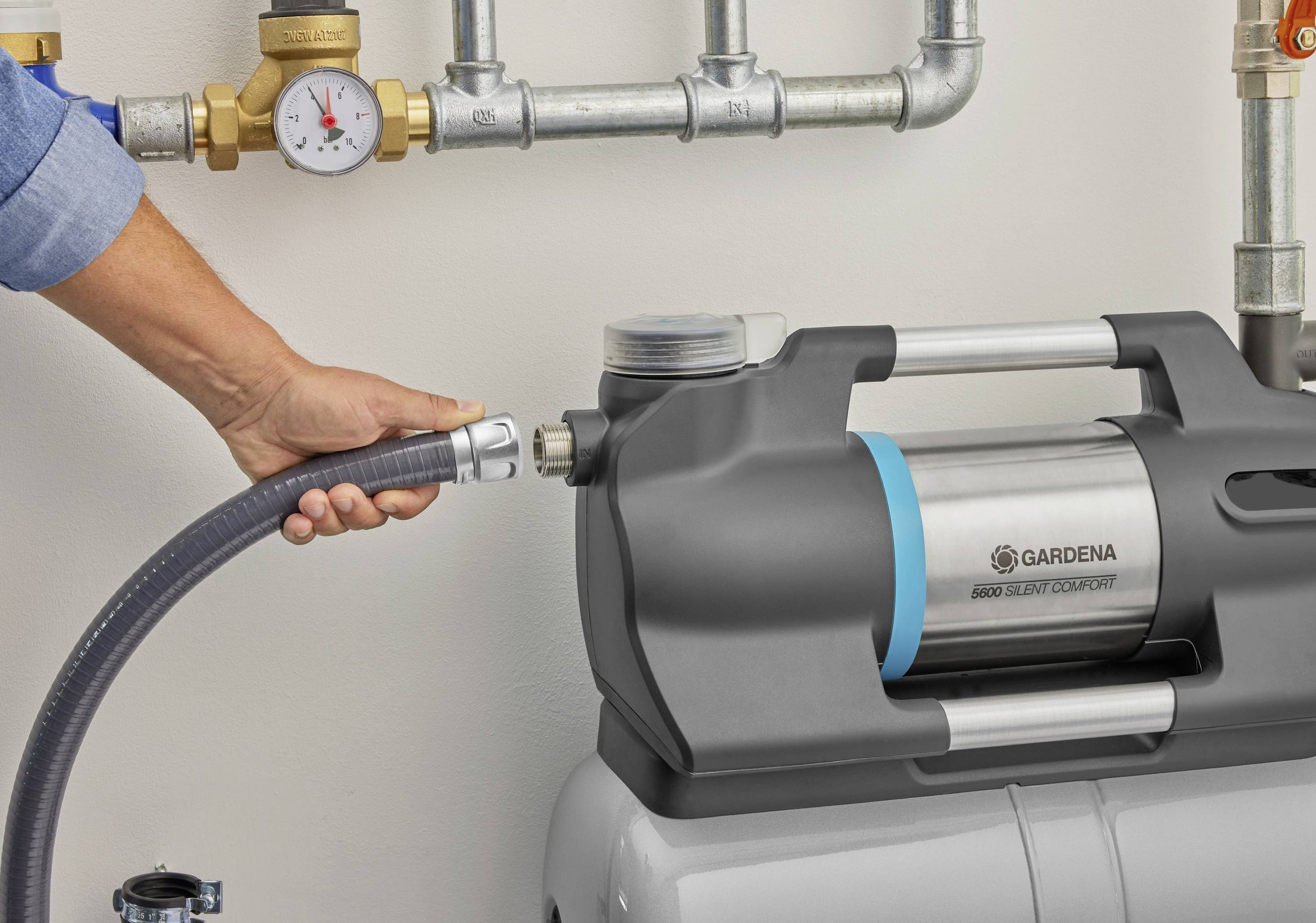 Osoba podłącza wąż ogrodowy do pompy wodnej Gardena. W tle widoczne są rury z manometrem.