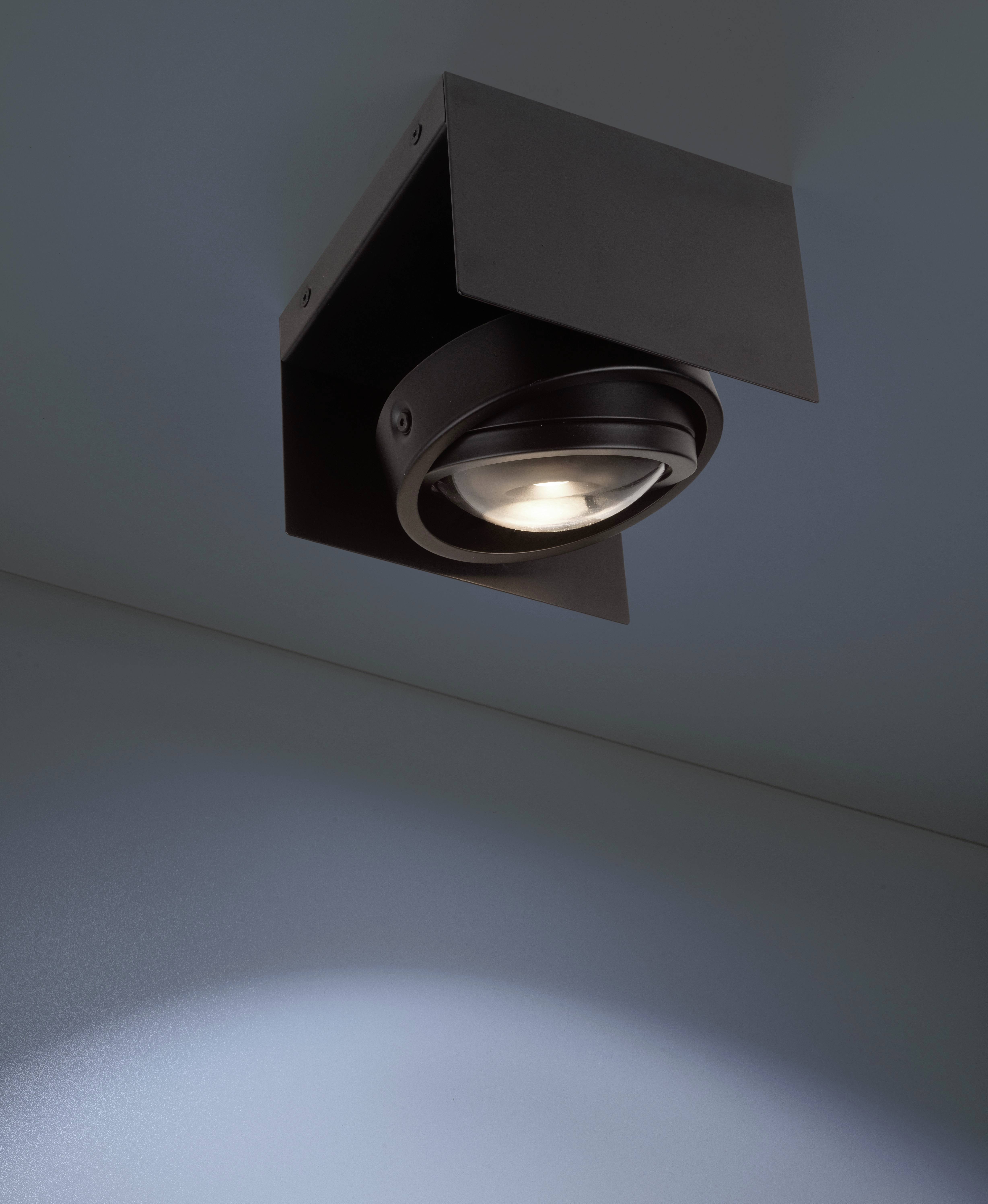 Ciemna lampa sufitowa, która świeci w dół i rzuca delikatne światło na białą ścianę.