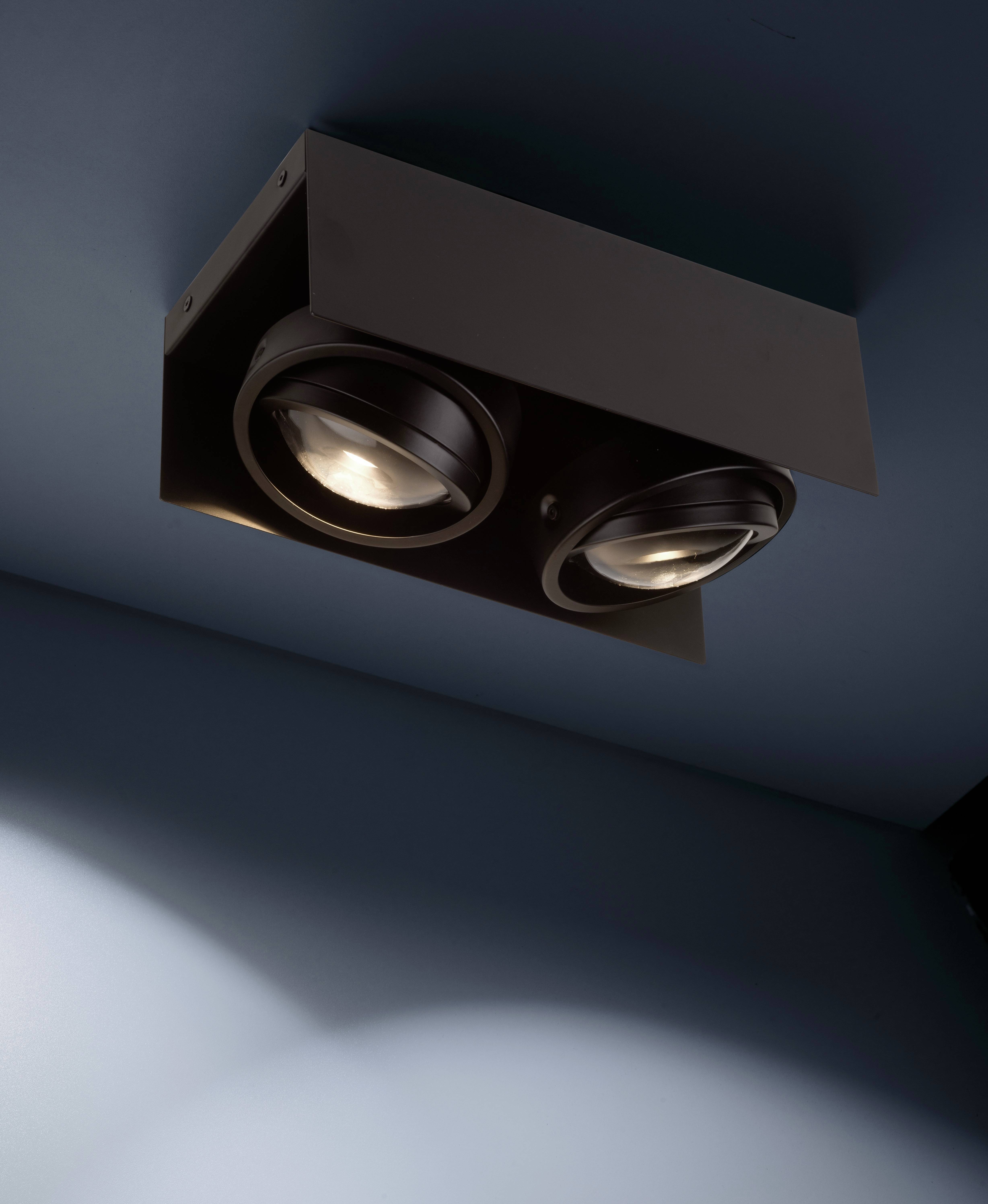 Nowoczesna lampa sufitowa z dwoma okrągłymi reflektorami zamontowanymi w ciemnej obudowie rzuca światło na niebieską ścianę.